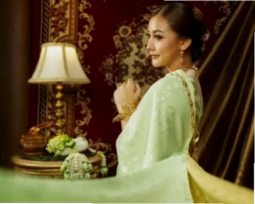 Phụ nữ mặc bộ trang phục Thái màu xanh nhạt, chắp tay, thể hiện sự trang nhã và duyên dáng trong khung cảnh hoa văn tinh xảo.