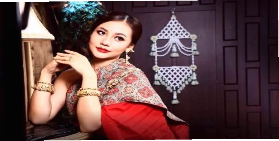 Quý cô mặc trang phục Thái màu đỏ vàng với mái tóc và trang điểm tinh tế, tạo dáng tay duyên dáng, mỉm cười quay đầu nhìn.