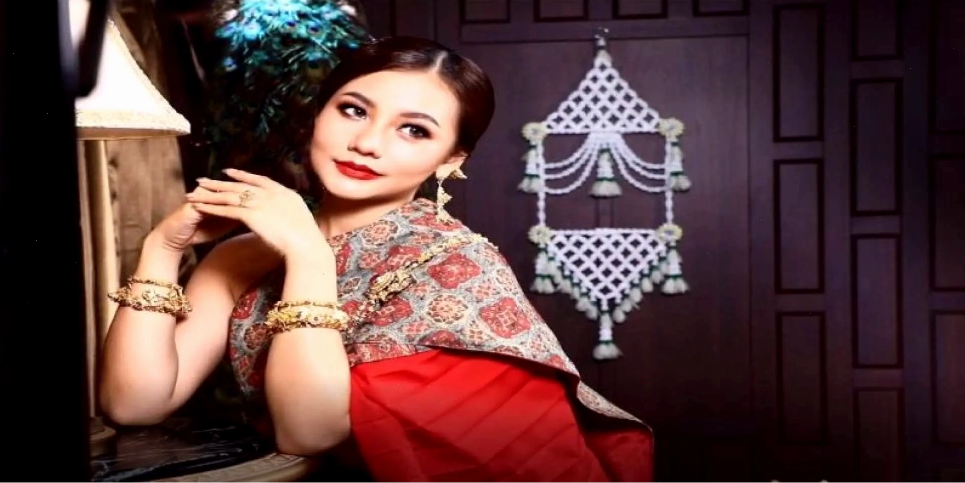 Quý cô mặc trang phục Thái màu đỏ vàng với mái tóc và trang điểm tinh tế, tạo dáng tay duyên dáng, mỉm cười quay đầu nhìn.