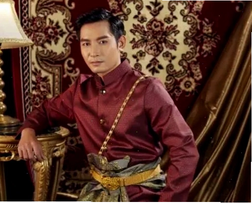 Quý ông mặc trang phục Thái màu đỏ rượu, tựa nhẹ vào bàn, thể hiện vẻ phong nhã trong khung cảnh sang trọng.
